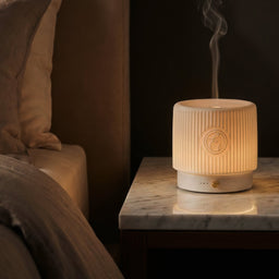 White ceramic diffuser emitting steam on a marble nightstand next to a bed.