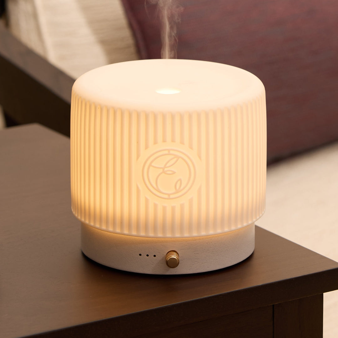 A cylindrical LED diffuser and humidifier with a ribbed white body, glowing warm yellow light, and a small gold button on the base, placed on a dark wooden table in a cozy indoor setting.