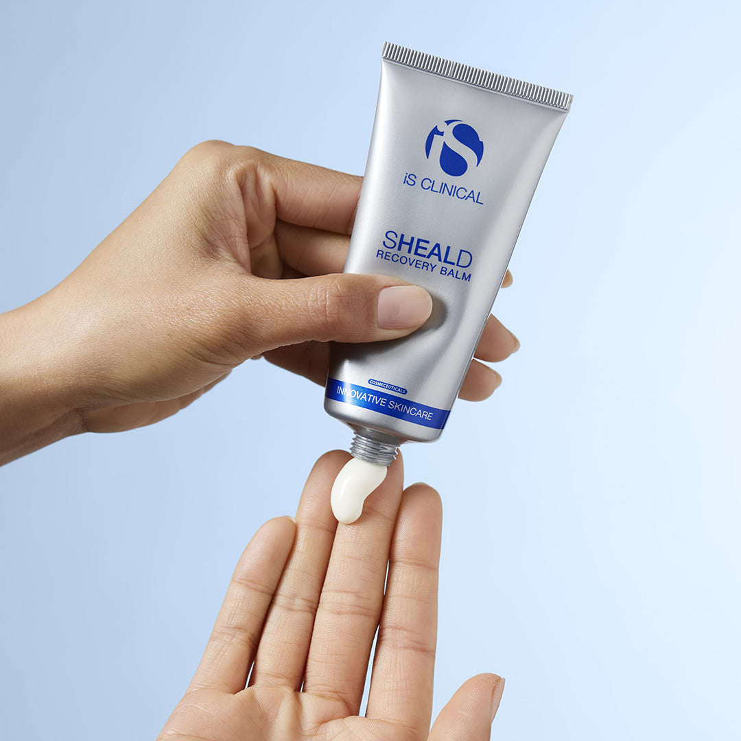 Hand holding a tube of IS Clinical SHEALD Recovery Balm against a light blue background
