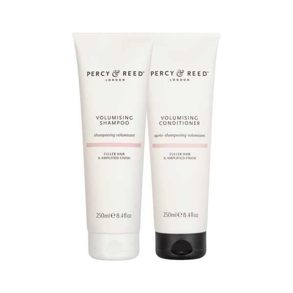 Two white tubes of Percy & Reed London haircare products on a white background: Volumising Shampoo with a pink accent band and Volumising Conditioner with a black flip-top cap.