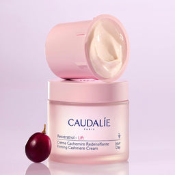 Caudalie Resveratrol - Lift Firming Cashmere Cream jar in pale pink, with the lid off and cream visible, alongside a dark red cranberry on a light lavender background.