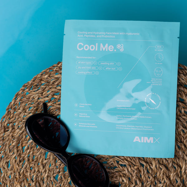 A light blue AIMX 'Cool Me' cooling face mask packet lies on a woven straw mat with black sunglasses, set against a solid turquoise background.