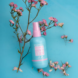 A light blue spray bottle of HairSkins Volume Growth Elixir with a pink cap, styled on a bright blue background with pink flowers.