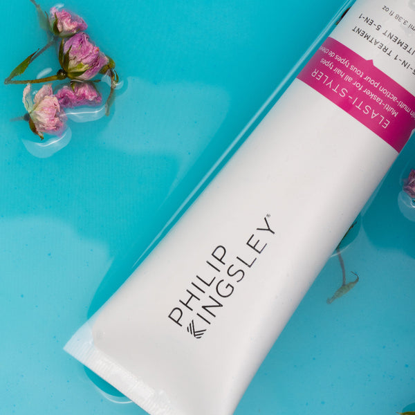 A white tube of Philip Kingsley Elast-Styleer treatment cream with a pink label, placed diagonally on a bright blue surface with small pink flowers floating around it.