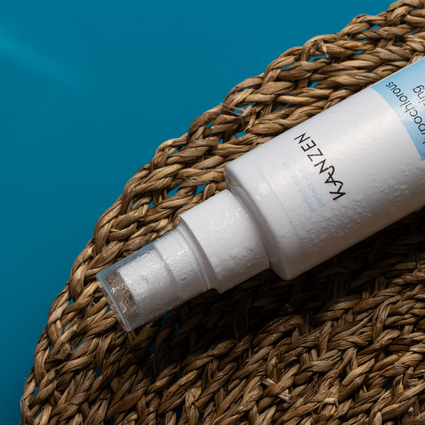 A white cylindrical spray bottle labeled KAVZEN Haircare Hair Reviver Spray, resting diagonally on a brown woven straw mat against a bright blue background.