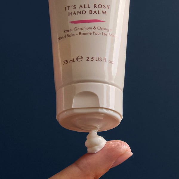 Hand applying 'It's All Rosy' hand balm to a finger against a dark background
