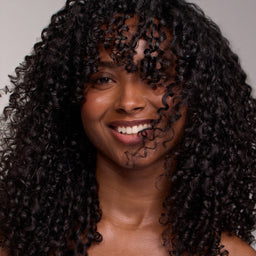 A close-up portrait of a smiling woman with voluminous, tightly curled black hair, shown against a plain light gray background.