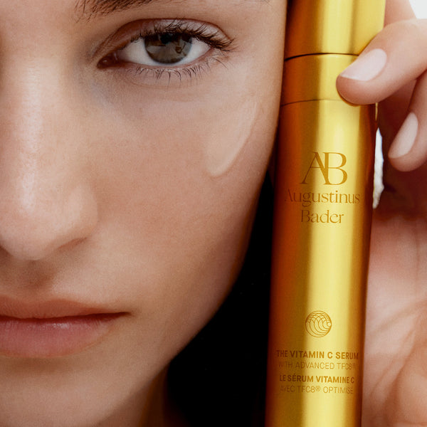 Close-up of a woman's face with a gold skincare serum bottle labeled 'Augustinus Bader' held close to her eye.