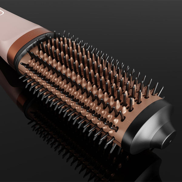 A close-up studio shot of a round hairbrush with a pale pink handle, bronze-toned bristles, and a dark metallic end, set against a glossy black background.