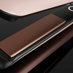 Close-up of a hair straightener with metallic rose gold and black surfaces, shown on a dark reflective background.