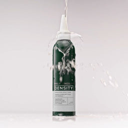 A single cylindrical bottle of DENSITY brand product stands upright on a white surface against a light gray studio background, with water splashing around it and a clear stream entering the top opening.