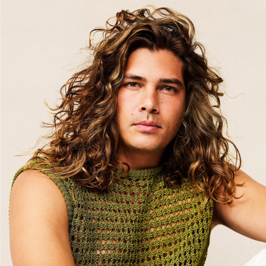 Man with long, wavy hair wearing a green sleeveless top against a beige background