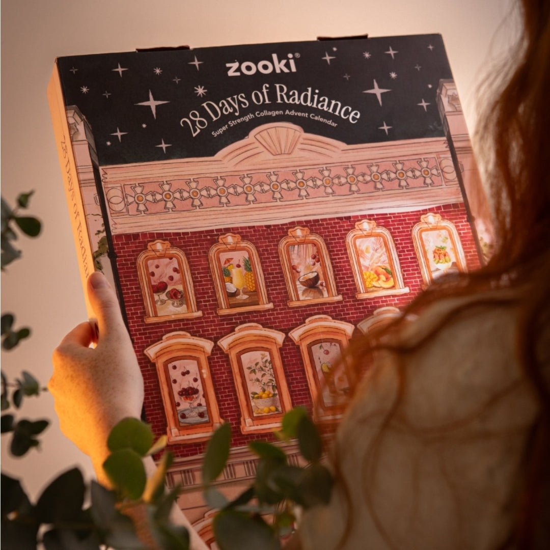A person holds a Zooki 28 Days of Radiance luxury chocolate advent calendar box. The box features a red brick building design with illustrated windows and a black starry top banner.