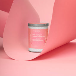 A cylindrical jar of Skin Collagen Support Superfood by The Glow Lab, featuring a pink label and silver lid, set against a minimalist pink background with draped paper.