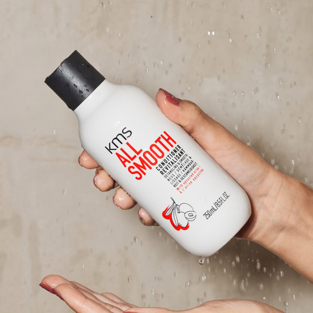 Hand holding a KMS All Smooth conditioner bottle against a shower background