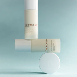 Three Omorovicza Balancing Moisturizer bottles in soft off-white and pale blue tones, arranged on a matching light blue background with one bottle standing upright, one lying horizontally, and a round cap placed in front.
