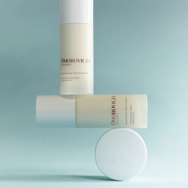 Three Omorovicza Balancing Moisturizer bottles in soft off-white and pale blue tones, arranged on a matching light blue background with one bottle standing upright, one lying horizontally, and a round cap placed in front.