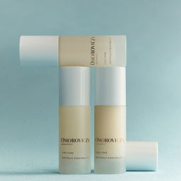 Three Omorovicza Ever Tone skincare bottles arranged in a studio setup against a soft light blue background. The bottles are frosted with pale beige liquid inside and have white caps, with one bottle lying horizontally behind the others.