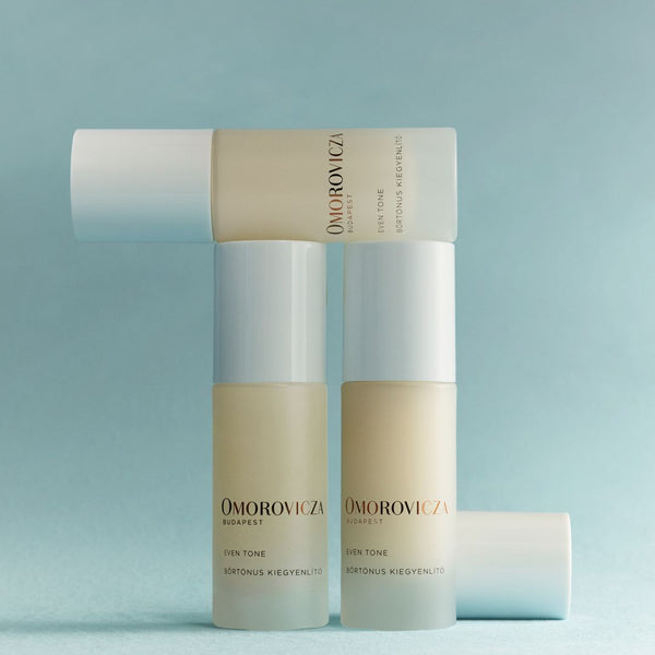 Three Omorovicza Ever Tone skincare bottles arranged in a studio setup against a soft light blue background. The bottles are frosted with pale beige liquid inside and have white caps, with one bottle lying horizontally behind the others.