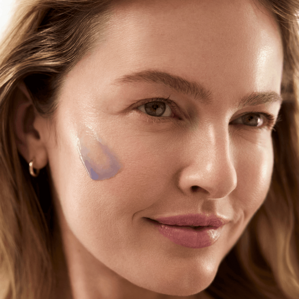 Close-up of a woman's face with a translucent blue-purple cosmetic swatch applied to her cheek, showing a glossy, dewy finish.