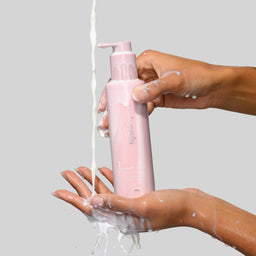 Pink bottle of shampoo being held over a hand with soap suds on a gray background