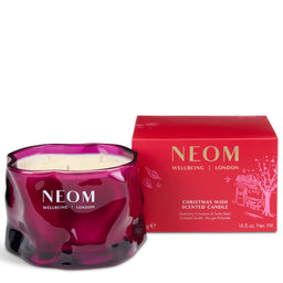A NEOM Wellbeing London 'Christmas Wish' scented candle in a wavy, translucent deep magenta glass jar, shown next to its red packaging box on a white background.
