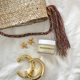 Wonderbalm jar with gold earrings and decorative bag on a textured white surface