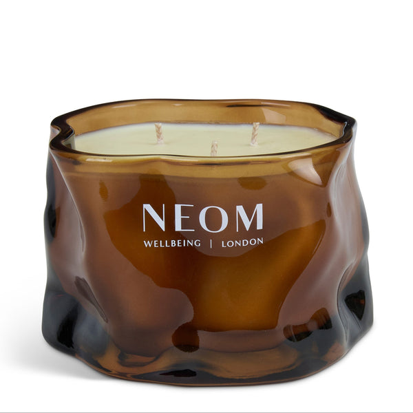 A NEOM Wellbeing London scented candle in an organic, wavy amber-brown glass container, featuring a cream-colored wax fill and white brand lettering on the front.