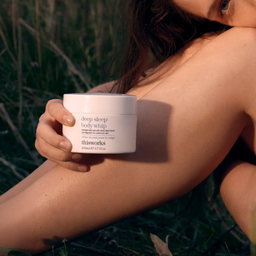 A person outdoors in grass holding a white jar of thisworks Deep Sleep Body Whip, with warm natural sunlight and soft focus on a bare shoulder.