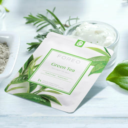 FOREEO Green Tea Purifying Mask in a white and green foil pouch, styled on a light blue wooden surface with bowls of ingredients, rosemary, and a green leaf.