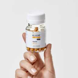 Hand holding a bottle of Heliocare capsules on a light gray background