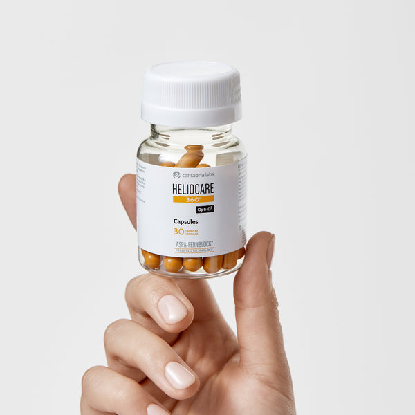 Hand holding a bottle of Heliocare capsules on a light gray background