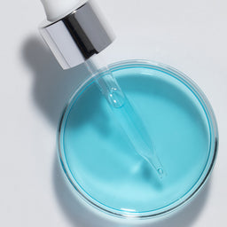 Blue liquid being dispensed from a dropper into a round container on a light gray background