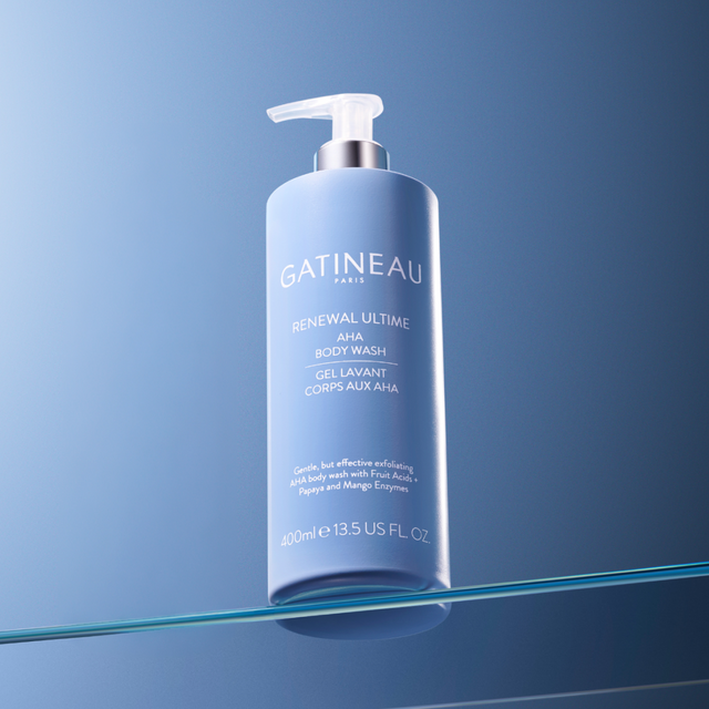A bottle of Gatineau Paris Renewal Ultime AHA Body Wash, 400ml, standing on a reflective glass surface against a blue gradient background.
