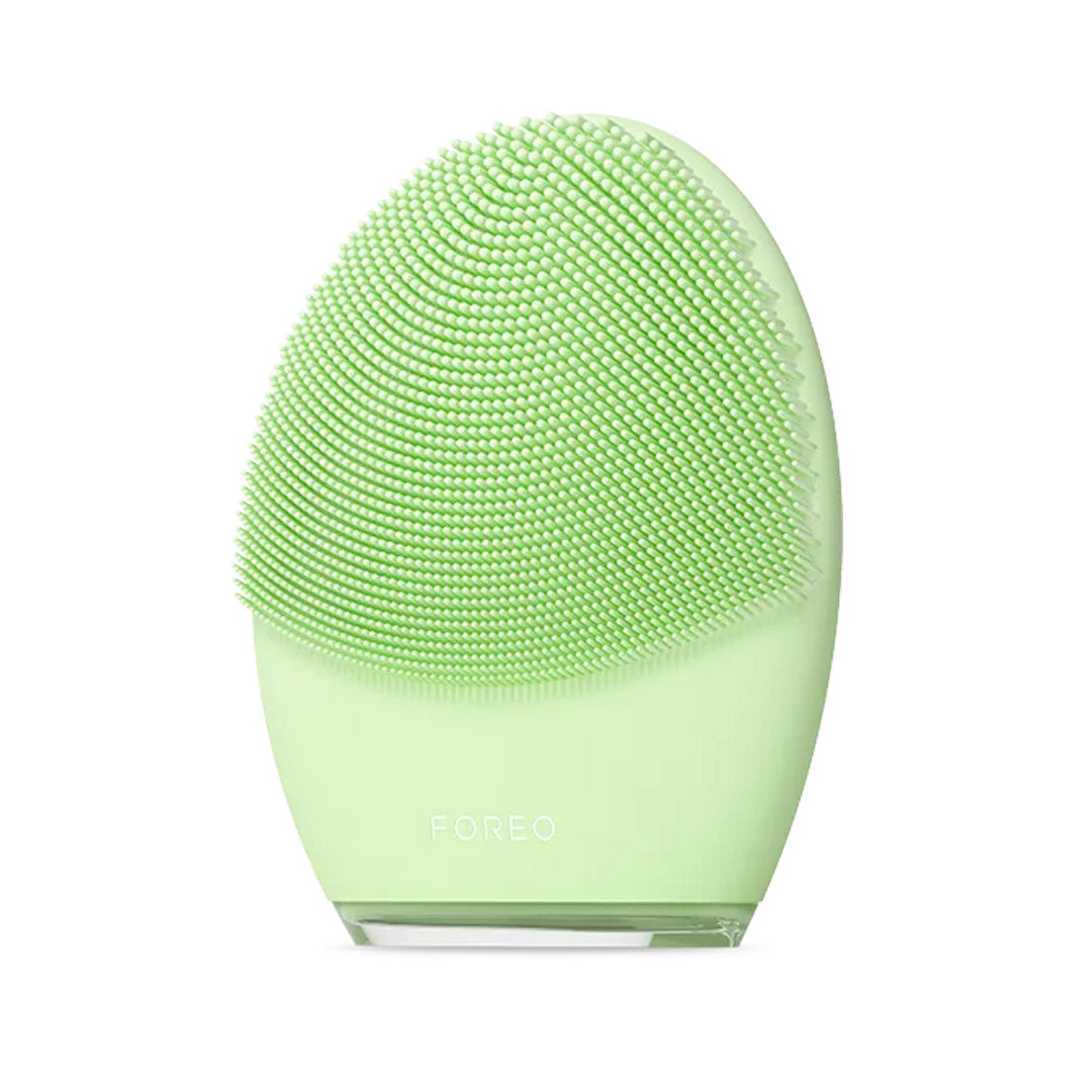 A FOREO Luna facial cleansing device in light green, featuring a curved silicone brush head and a matching green base with the FOREO logo, set against a plain white background.