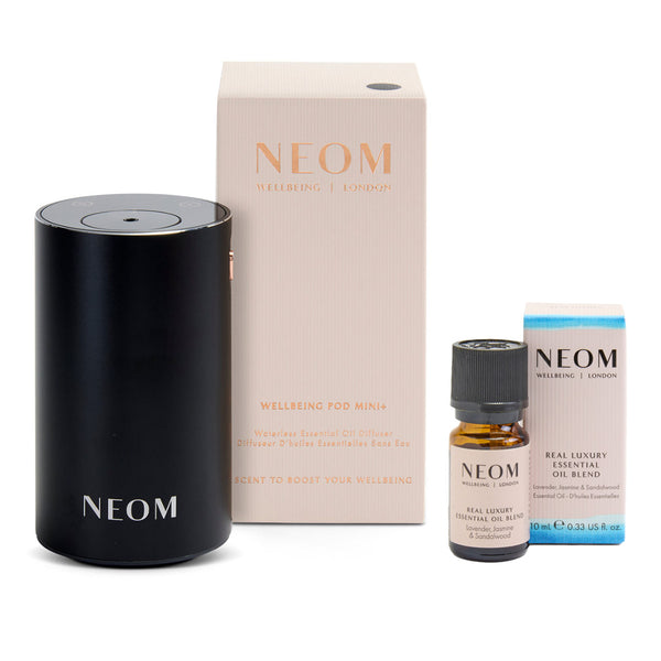 NEOM Welcoming Pod Mini diffuser set on a white background, including the matte black cylindrical diffuser, its cream-colored packaging box, a small amber essential oil bottle, and a blue-accented product box.