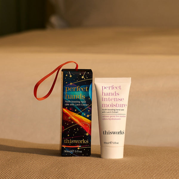 A tube of Thisworks Perfect Hands Intense Moisture hand care cream stands next to its dark blue packaging box with a red ribbon handle, set on a textured beige fabric surface.