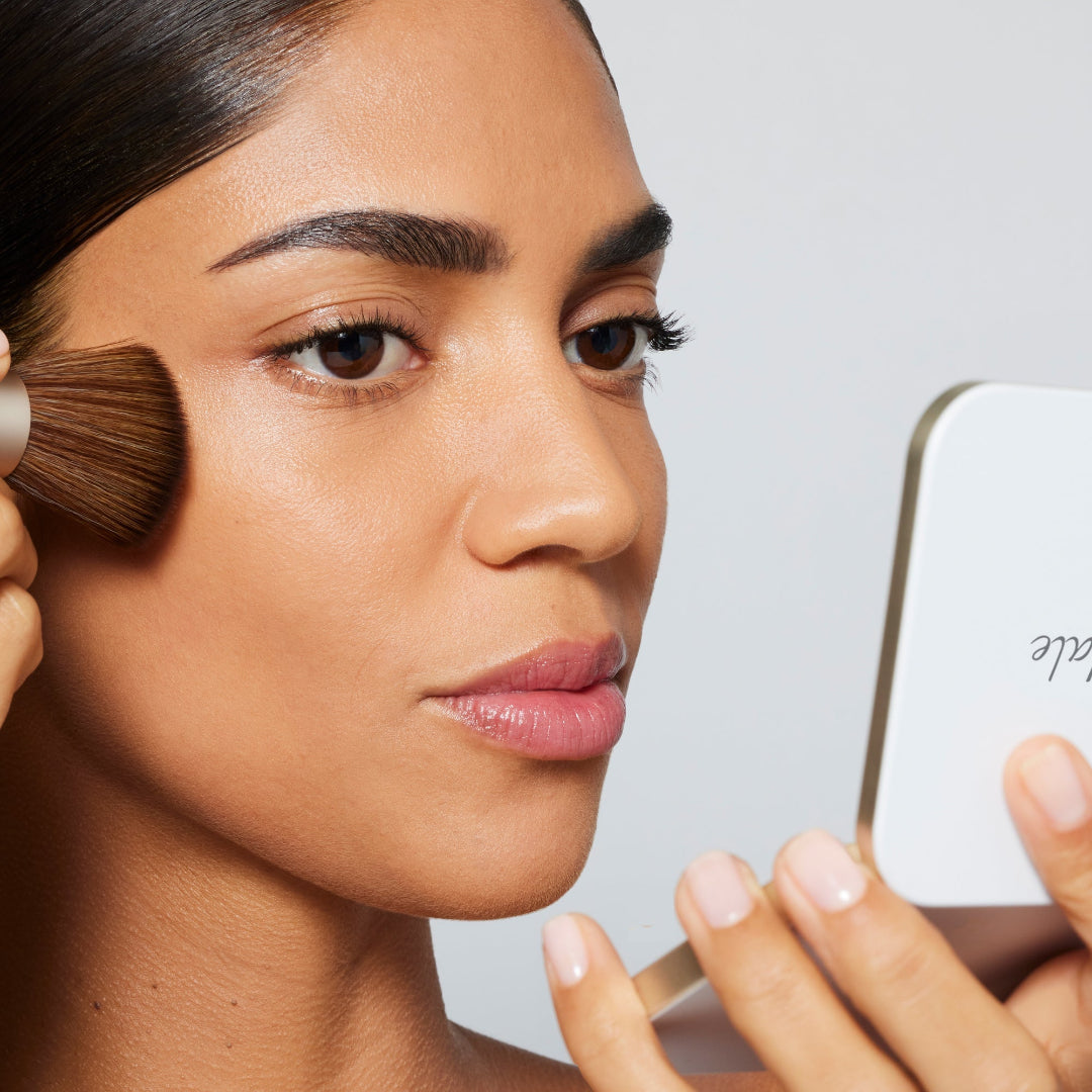 Woman applying makeup with a brush and looking at a phone, against a neutral background