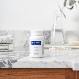 A white bottle of Pure Encapsulations Biotin 8 mg dietary supplement, set on a white marble countertop with a blurred bathroom background.