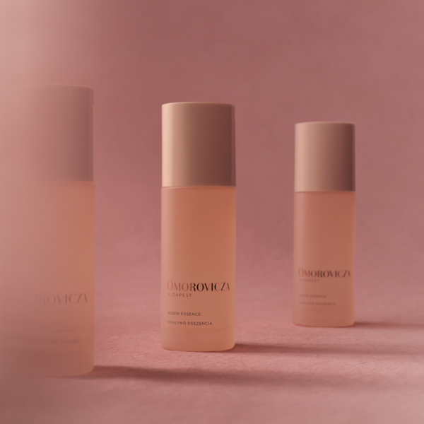Three Omorovicza Capsule skincare bottles with pale peach-pink liquid and beige caps, arranged on a soft pink background with a central bottle in sharp focus and blurred bottles on either side.