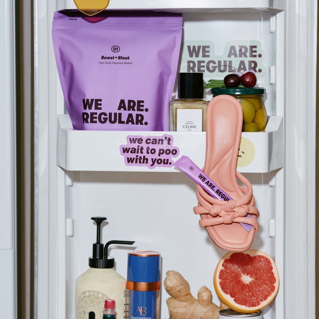 A styled refrigerator door scene with multiple props and text-based branding. The most salient sold item appears to be the pink slip-on shoe with a purple strap hanging on the middle shelf, surrounded by other objects including a purple Sweet Rusal bag, a perfume bottle, olives, ginger, a grapefruit slice, and Perello canned soda.