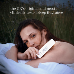 A woman lying on a white bed outdoors at dusk, holding a white rectangular sleep fragrance diffuser or perfume bottle against her shoulder. Tall grasses and a dark blue sky are blurred in the background, with white text at the top reading, 'the UK's original and most clinically tested sleep fragrance'.
