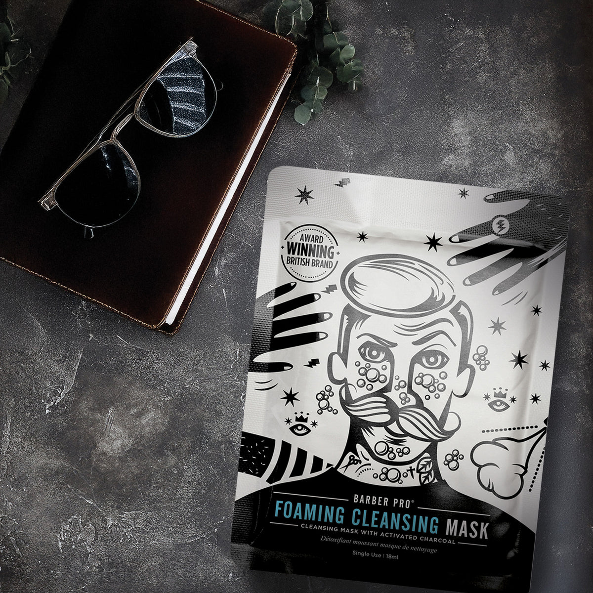 Foaming cleansing mask packaging with a stylized face design on a dark surface with sunglasses and a book.