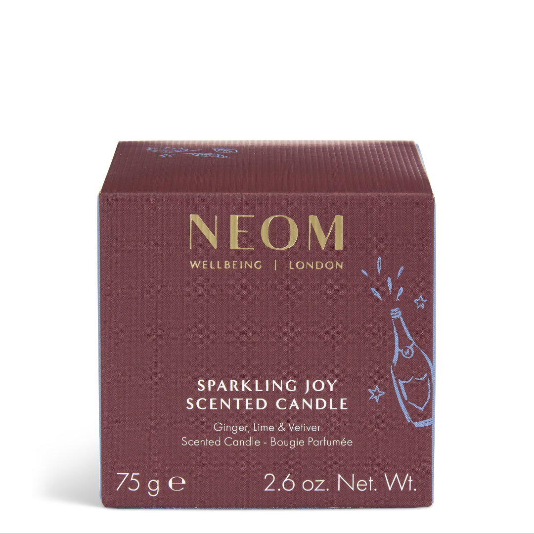 A square NEOM Wellbeing London 'Sparkling Joy' scented candle box in a deep burgundy color with gold lettering and a light blue champagne bottle illustration, set against a plain white background.