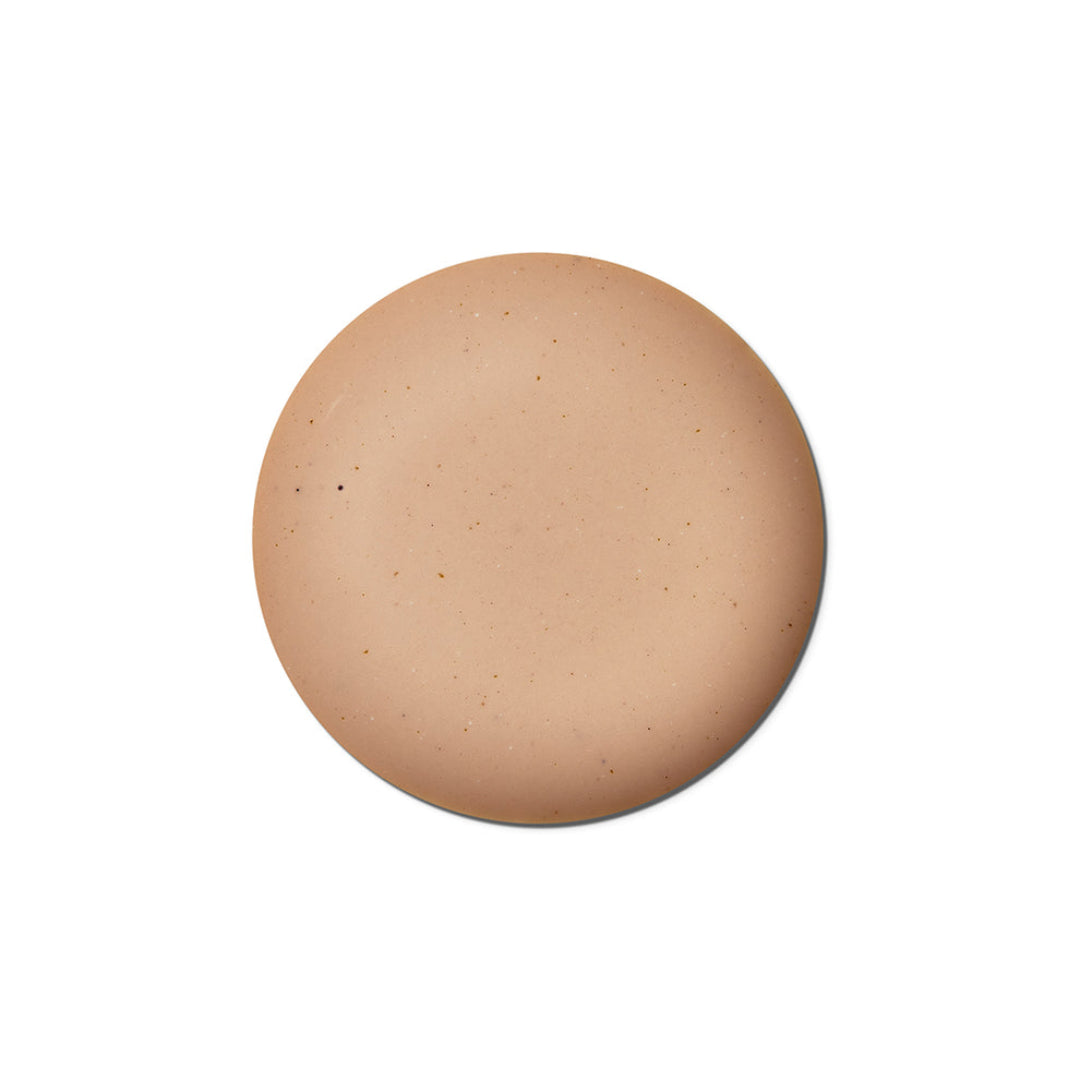 A circular makeup swatch showing a warm, light tan skin tone with subtle speckles, centered on a solid white background.