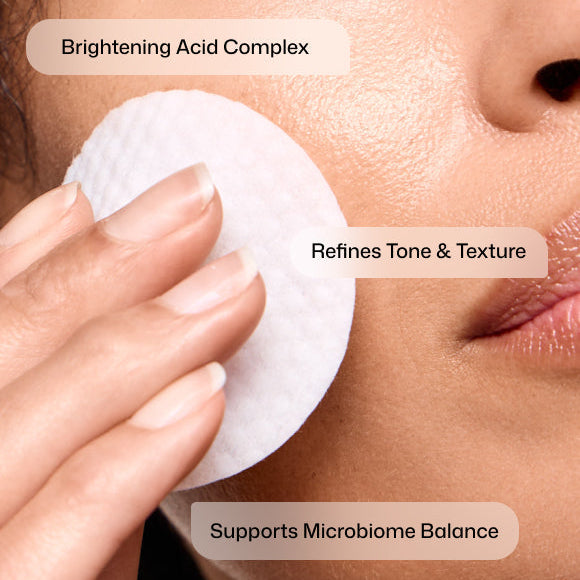 Close-up of a person's face and hand using a white round cotton pad to apply a brightening acid complex, with text overlays stating 'Brightening Acid Complex', 'Refines Tone & Texture', and 'Supports Microbiome Balance'.
