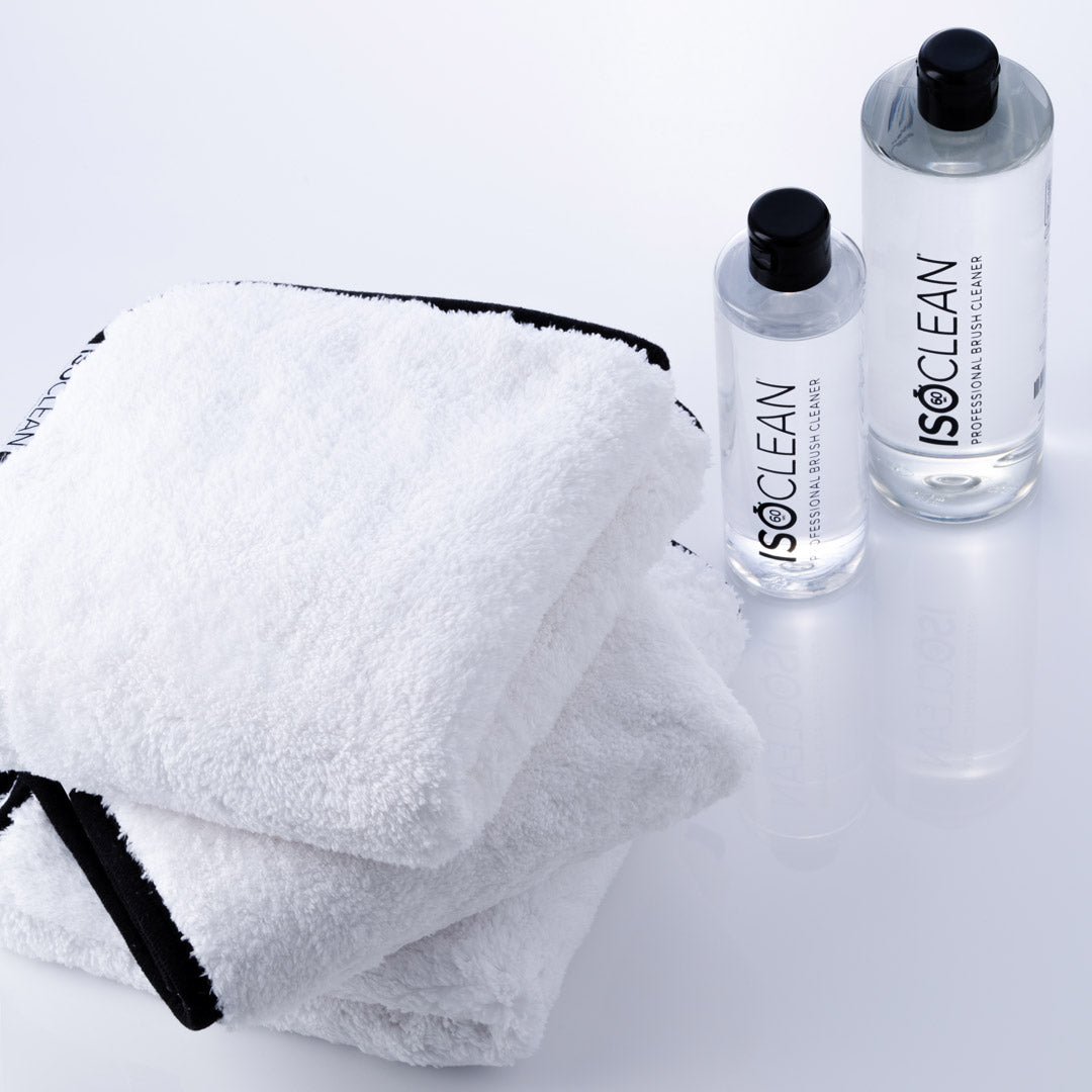 A folded white towel with black trim sits beside two clear bottles of ISOCLEAN professional basic cleaner on a clean white background.