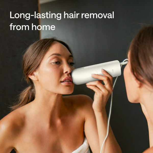 A woman uses a white handheld hair removal device on her upper chest while looking into a mirror. Text at the top reads 'Long-lasting hair removal from home.