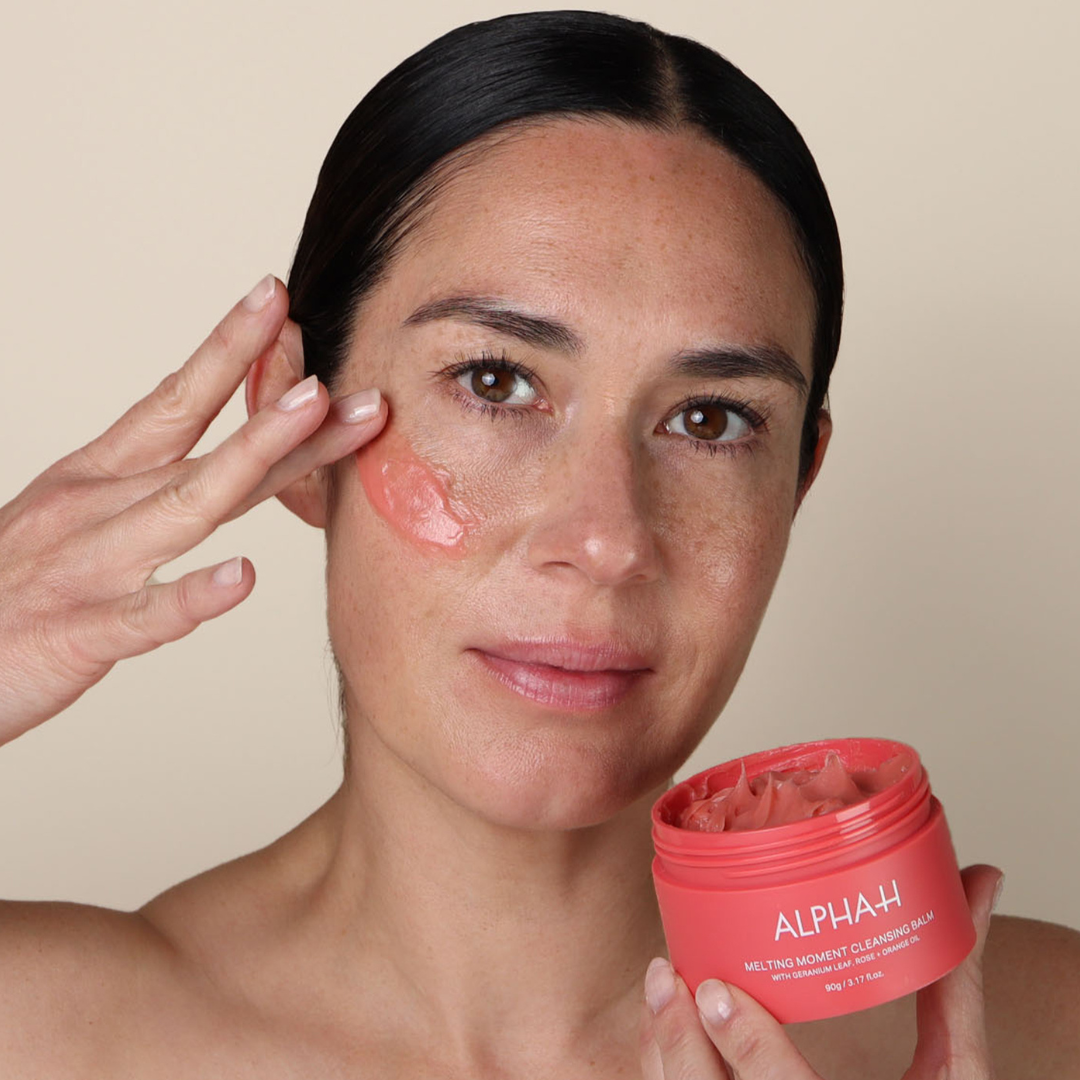 Woman applying a pink cream to her face with a container of the same cream in front of her.
