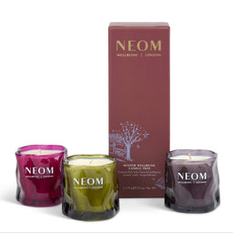 Three NEOM Wellbeing candles in textured glass jars with a tall mauve box behind them on a white background. The left candle is magenta, the center is olive green, and the right is dark purple, all labeled NEOM Wellbeing London.